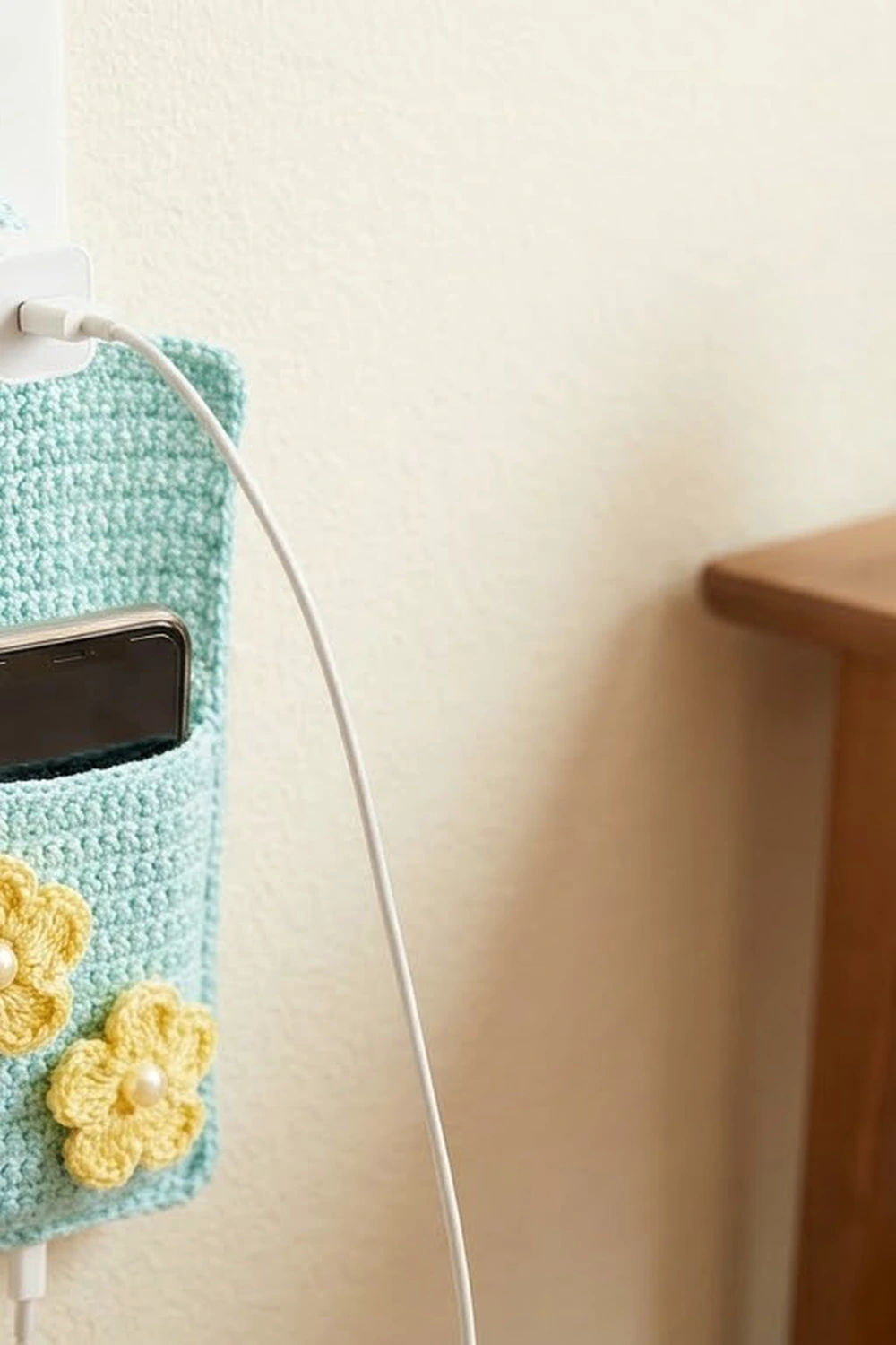 A seafoam crochet phone pouch with yellow flower appliqués holds a charging smartphone against a neutral wall.