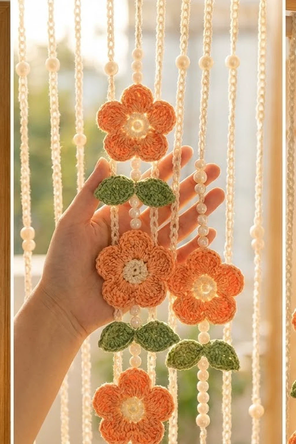 Hand holding a crocheted flower mobile with orange flowers, green leaves, and beaded cream strands backlit by a sunny window.