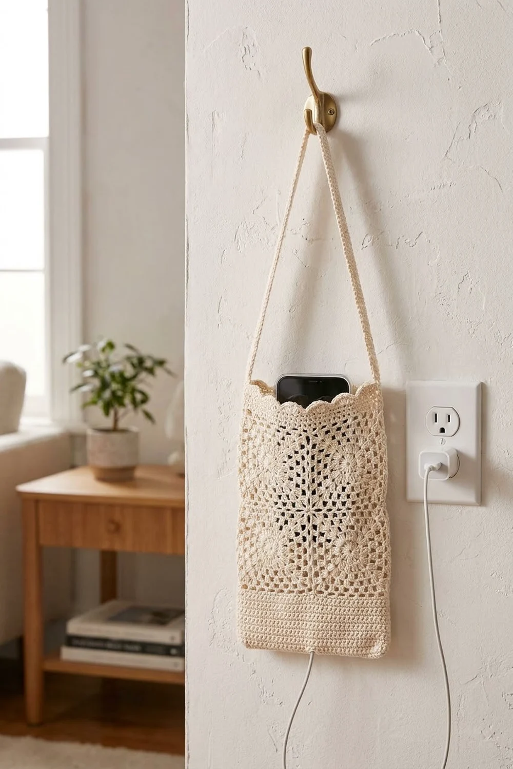 Cream lace-patterned crochet phone holder hanging from brass hook on white wall, containing a black smartphone with charging cable.