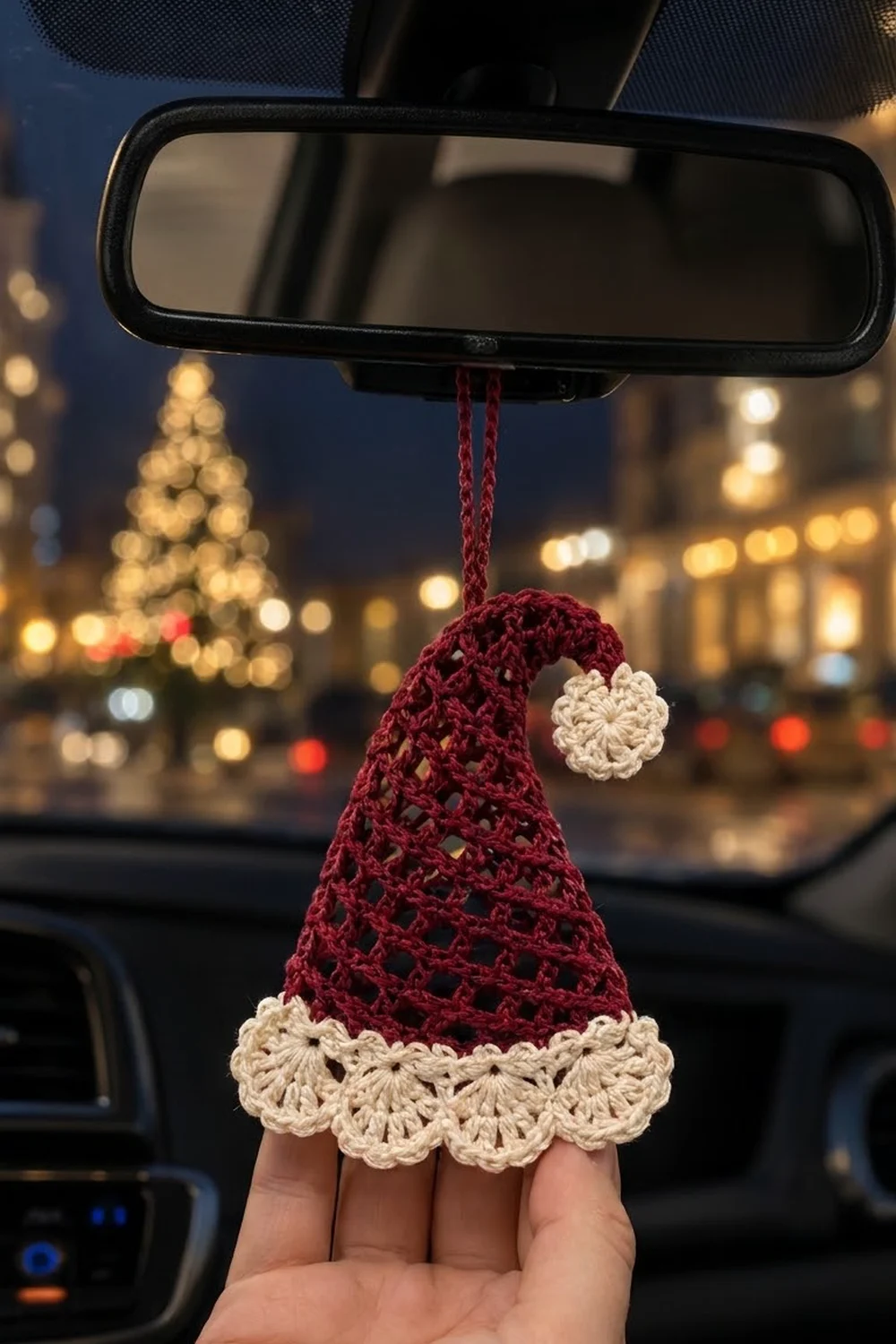Burgundy and cream crocheted Santa hat ornament hanging from rearview mirror with blurred holiday city lights and Christmas tree background.