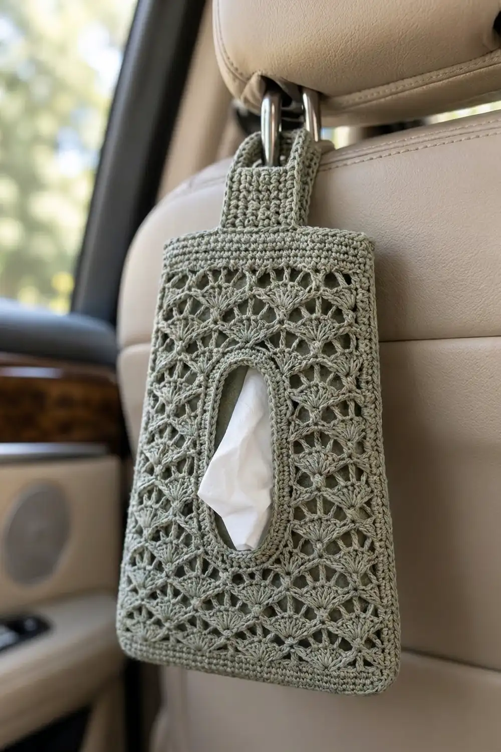 Sage-green crocheted tissue box holder with lace pattern hanging from a car headrest using metal hooks.