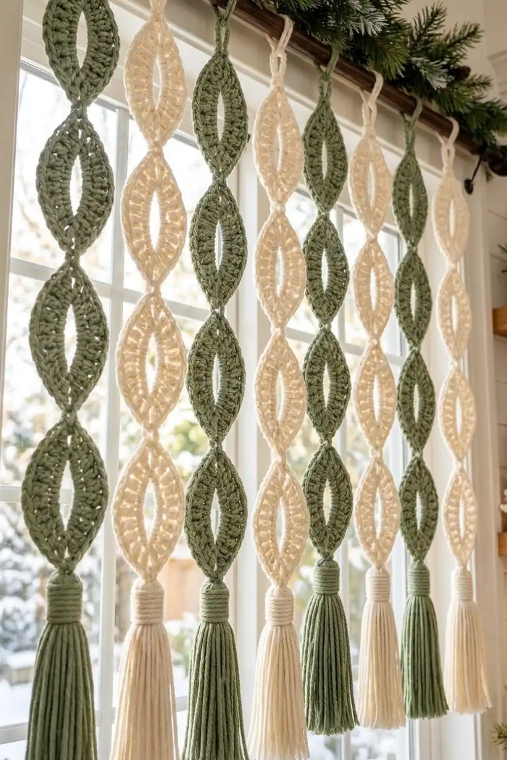 Crocheted leaf ornaments in sage green and cream hang from a window with evergreen garland above.