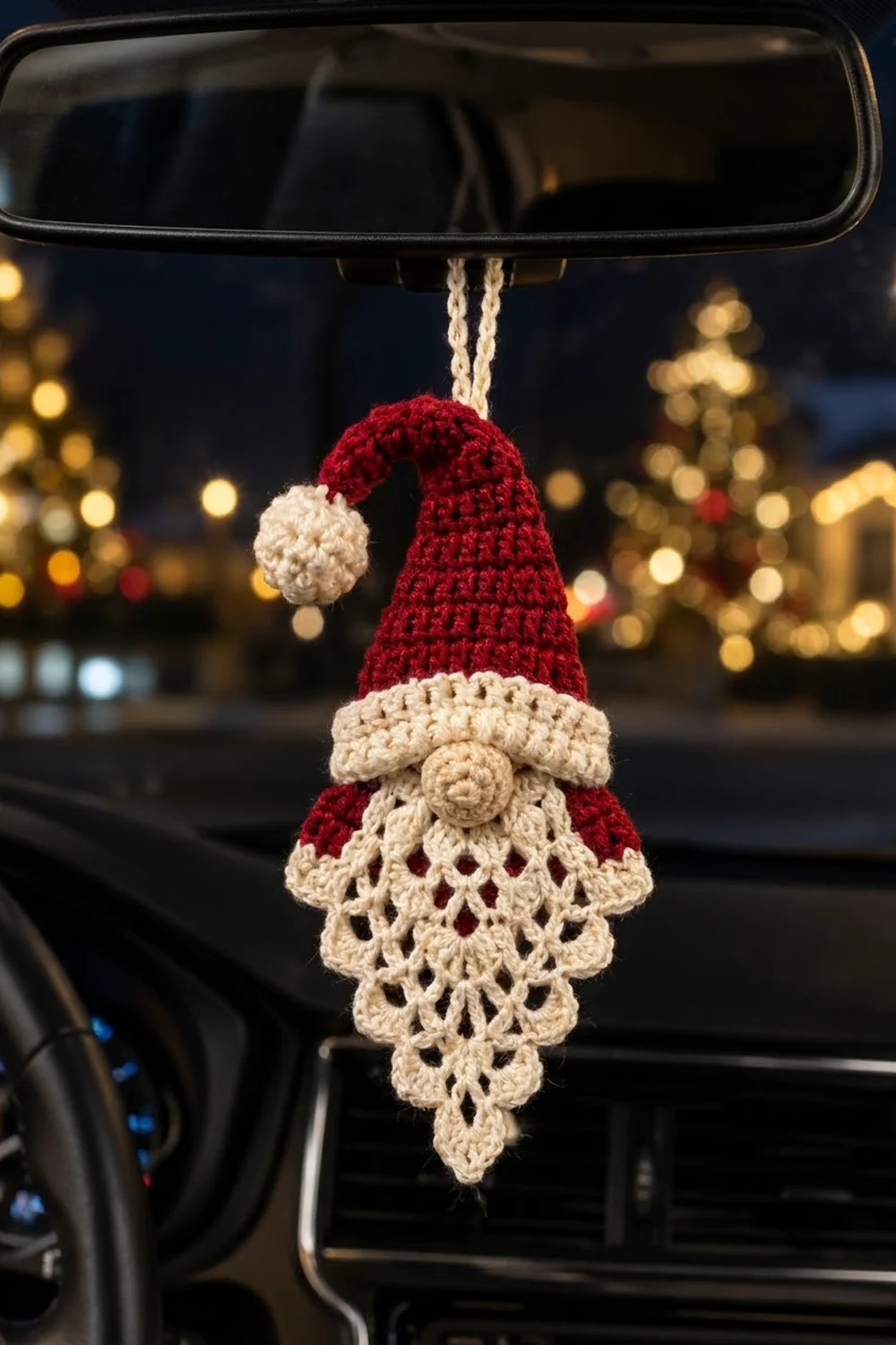 Crocheted red and cream gnome with lacy beard hanging from car rearview mirror against blurred holiday city lights.