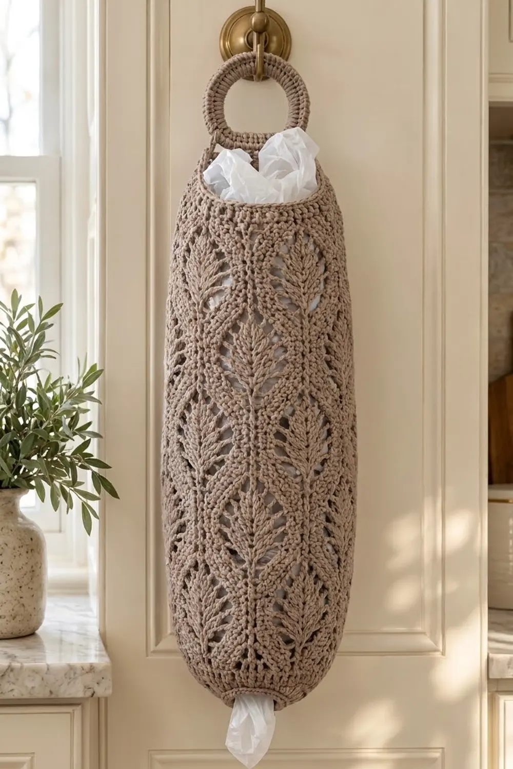 Taupe crocheted hanging organizer with delicate leaf pattern holds white tissues against cream wall.