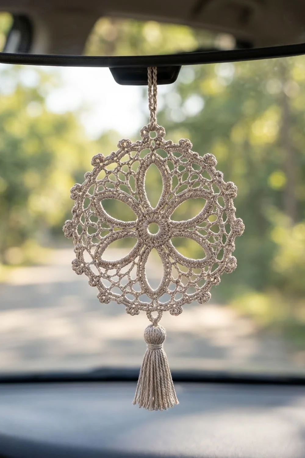 Delicate tan crocheted ornament with lace pattern and tassel hanging from a car mirror overlooking a blurred garden.