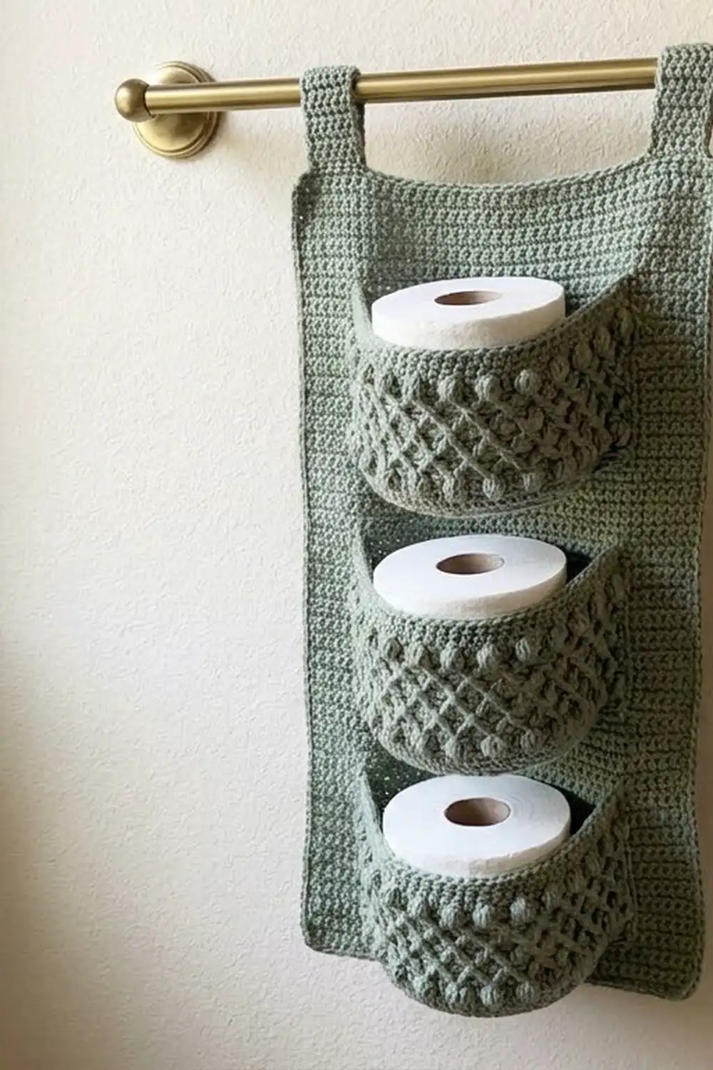 Sage green crocheted toilet paper holder with three pockets and decorative stitching, mounted on a brass rod against white wall.