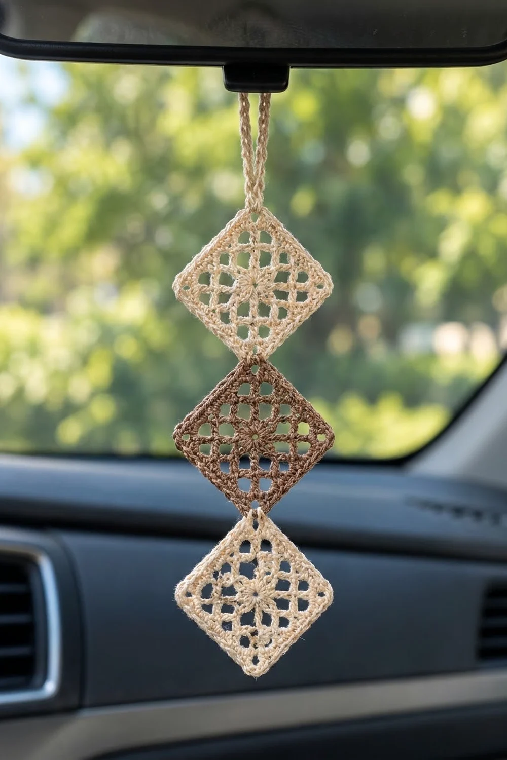 Beige and brown crocheted diamond-shaped air freshener hanging from car's rearview mirror with green outdoor background.