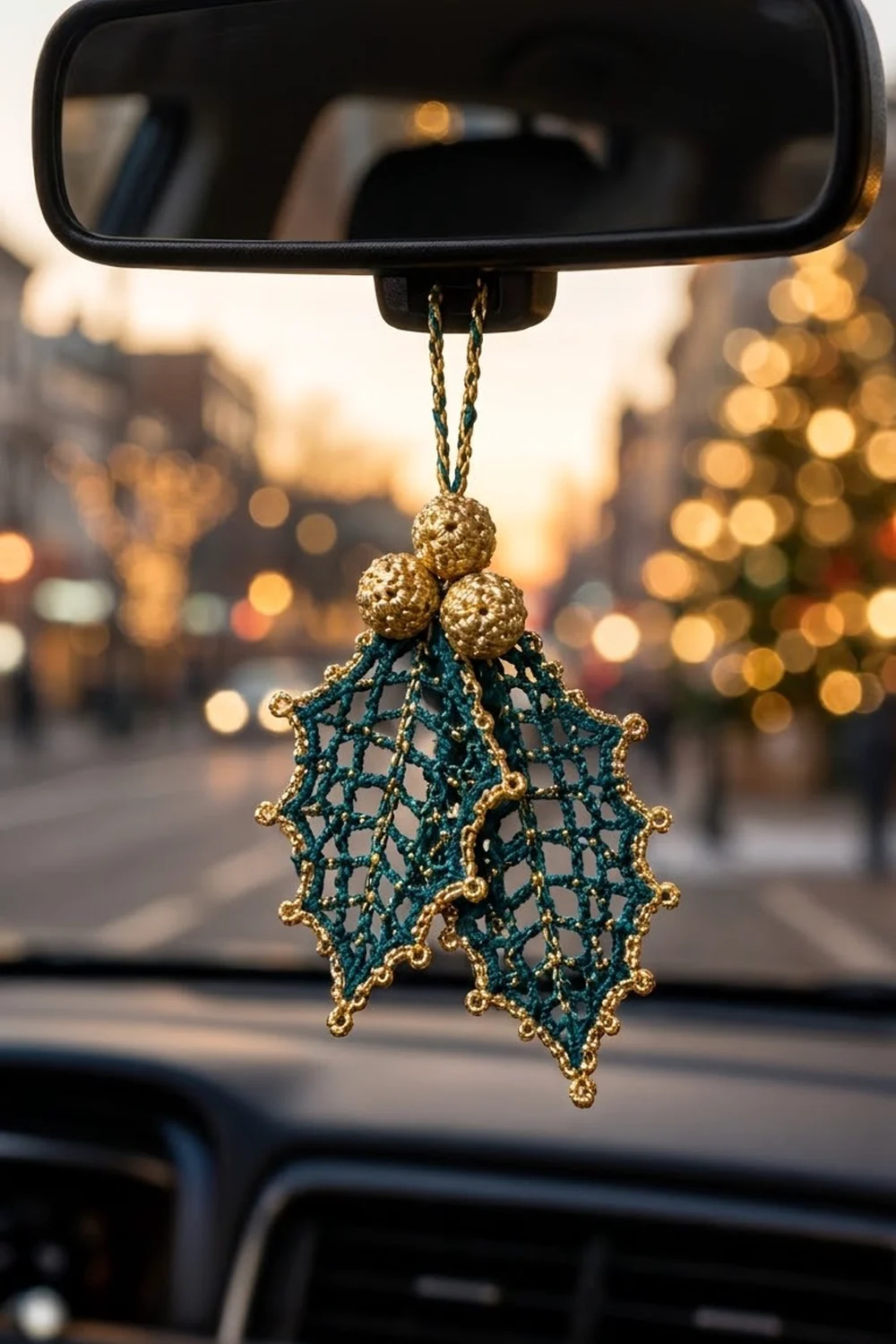 Crocheted teal-and-gold leaf ornament with beaded top hanging from a car rearview mirror against a warm-lit blurred street.