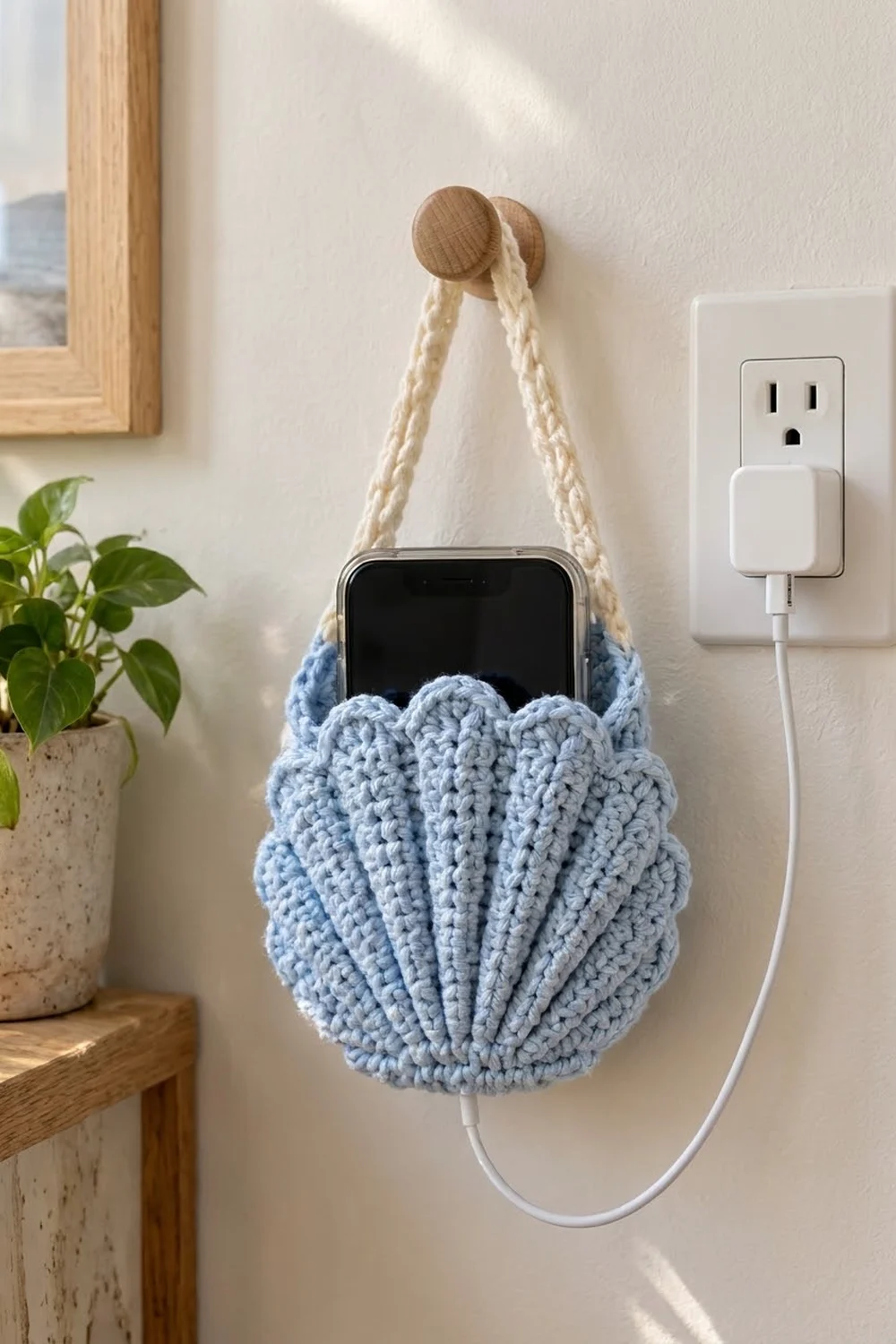 Light blue crocheted shell-shaped phone holder with rope strap hanging on white wall next to outlet.