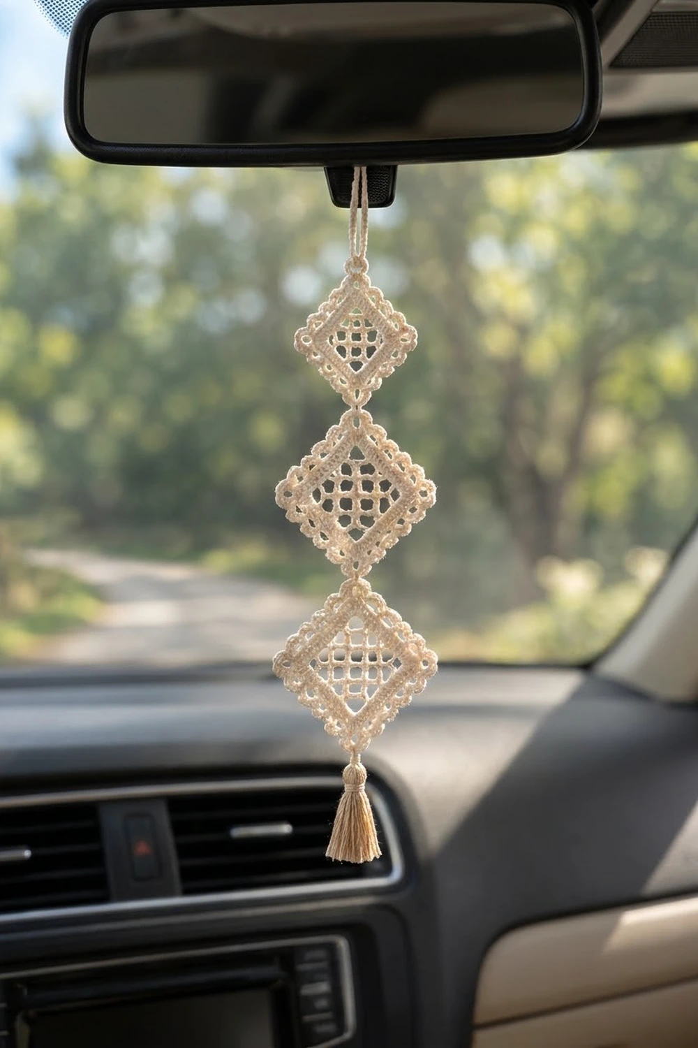 Beige crocheted three-diamond rearview mirror charm with openwork lattice pattern and tassel, hanging in sunny car with blurred green scenery.