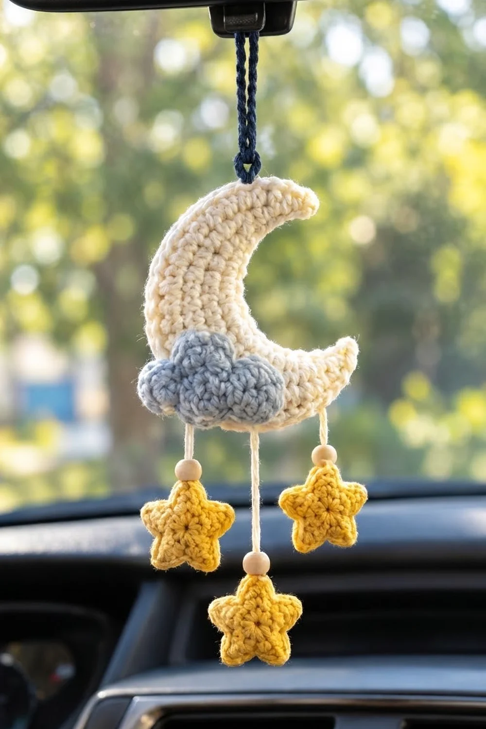 Cream and gray crocheted moon with hanging yellow stars, suspended from a car rearview mirror with blurred green background.