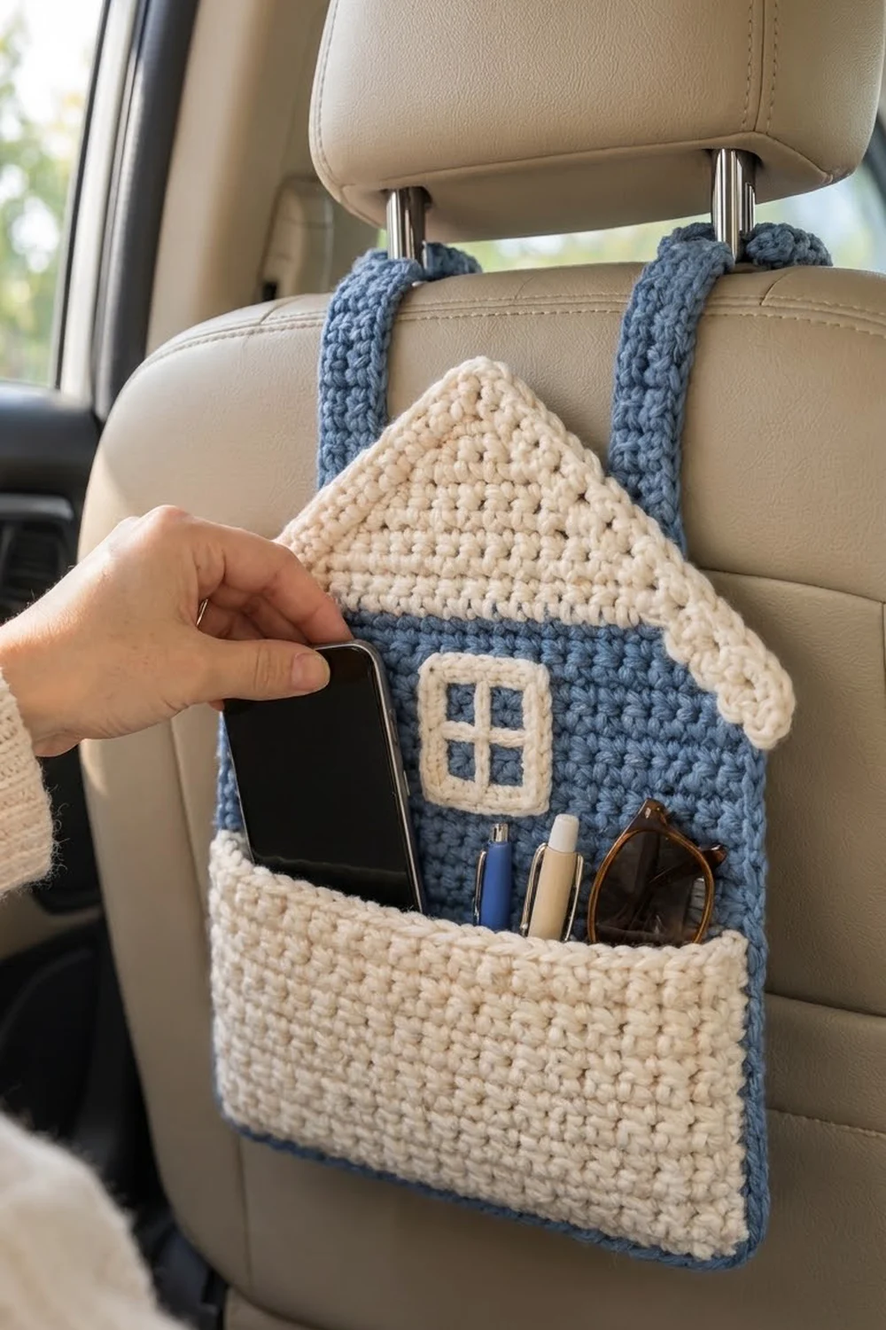 Blue and cream crocheted house organizer hanging from car seat headrest, displaying phone and accessories.