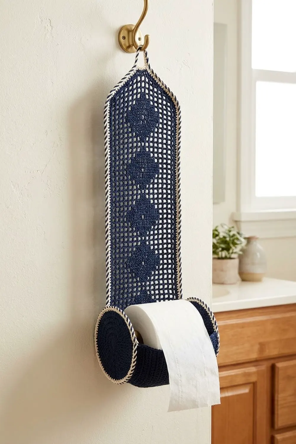 Navy and cream crocheted toilet paper dispenser with geometric pattern hanging on brass bathroom hook.