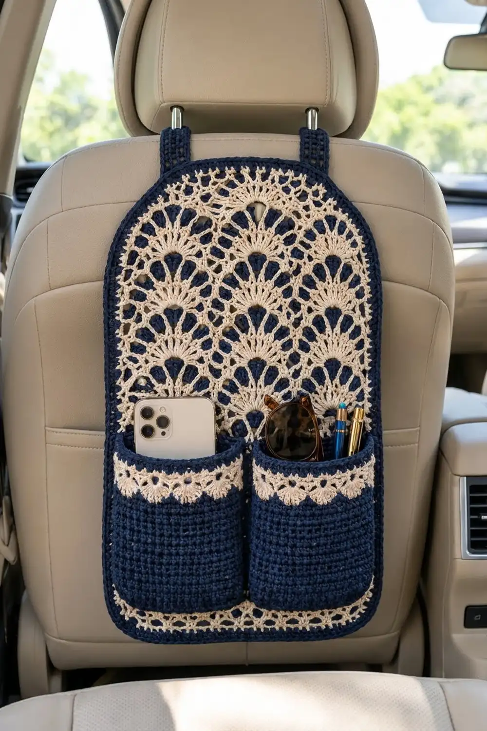 Navy crocheted car seat organizer with cream lace panels, holding a phone and accessories on a beige leather car seat.