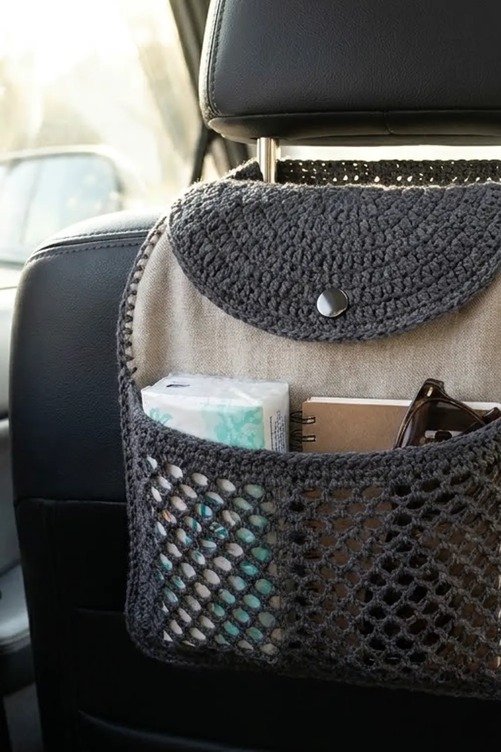 Charcoal gray crocheted car organizer with cream backing hanging from a car seat headrest, holding tissues and personal items.
