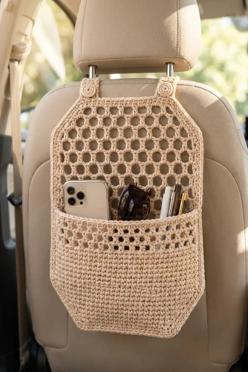 Cream crocheted car seat organizer with lacy lattice top and storage pocket holding a phone, sunglasses, and pens.