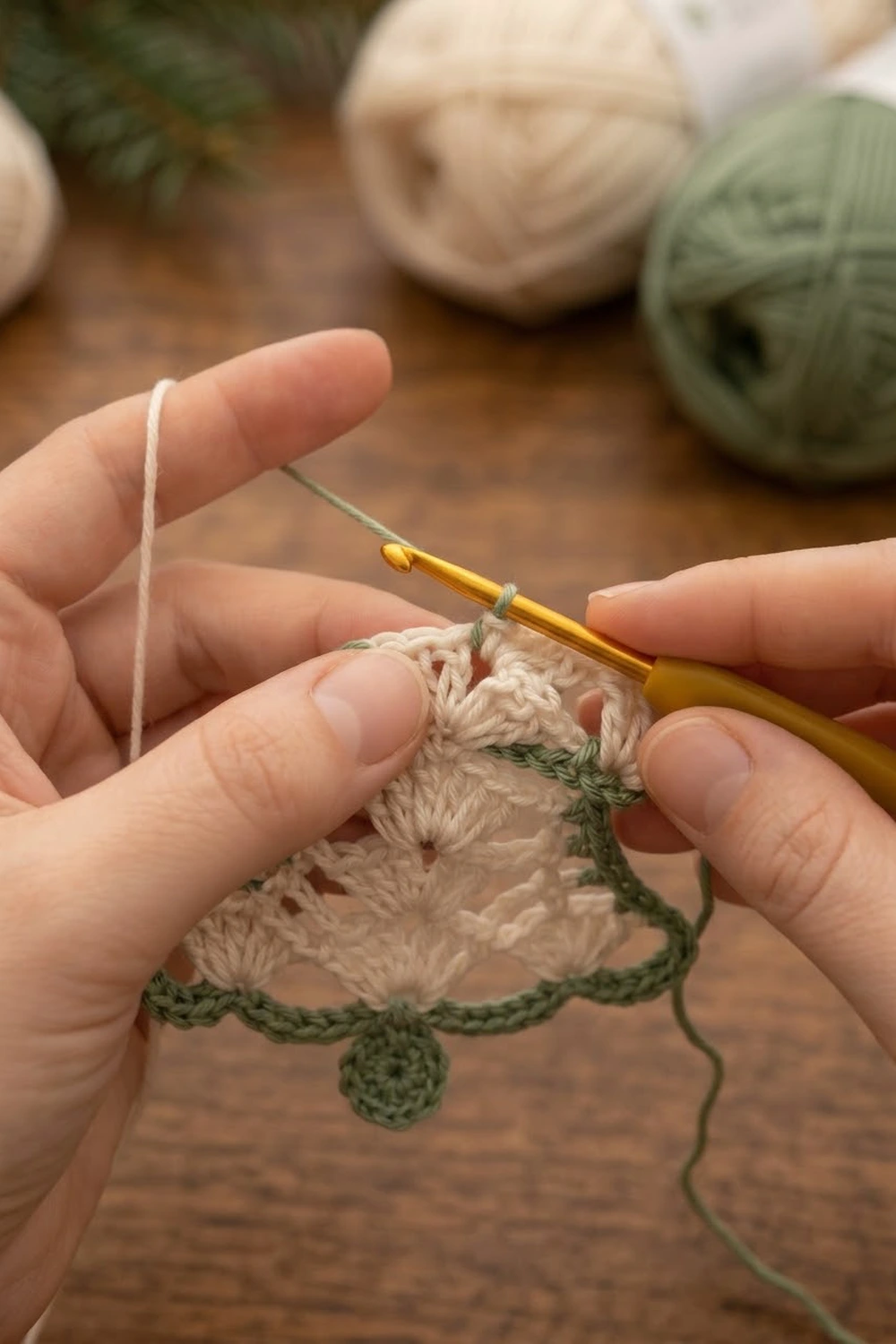 Hands crocheting a beige and green decorative piece with a golden hook on a wooden surface.