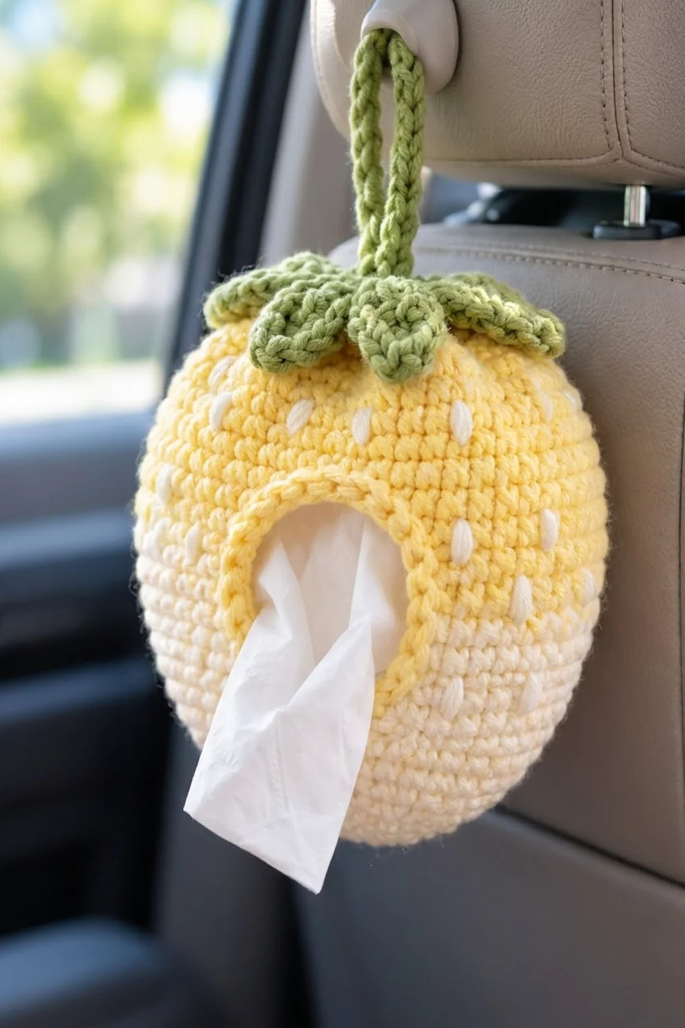 Yellow crocheted lemon-shaped tissue holder with green stem hanging from a car's sun visor dispensing tissue.