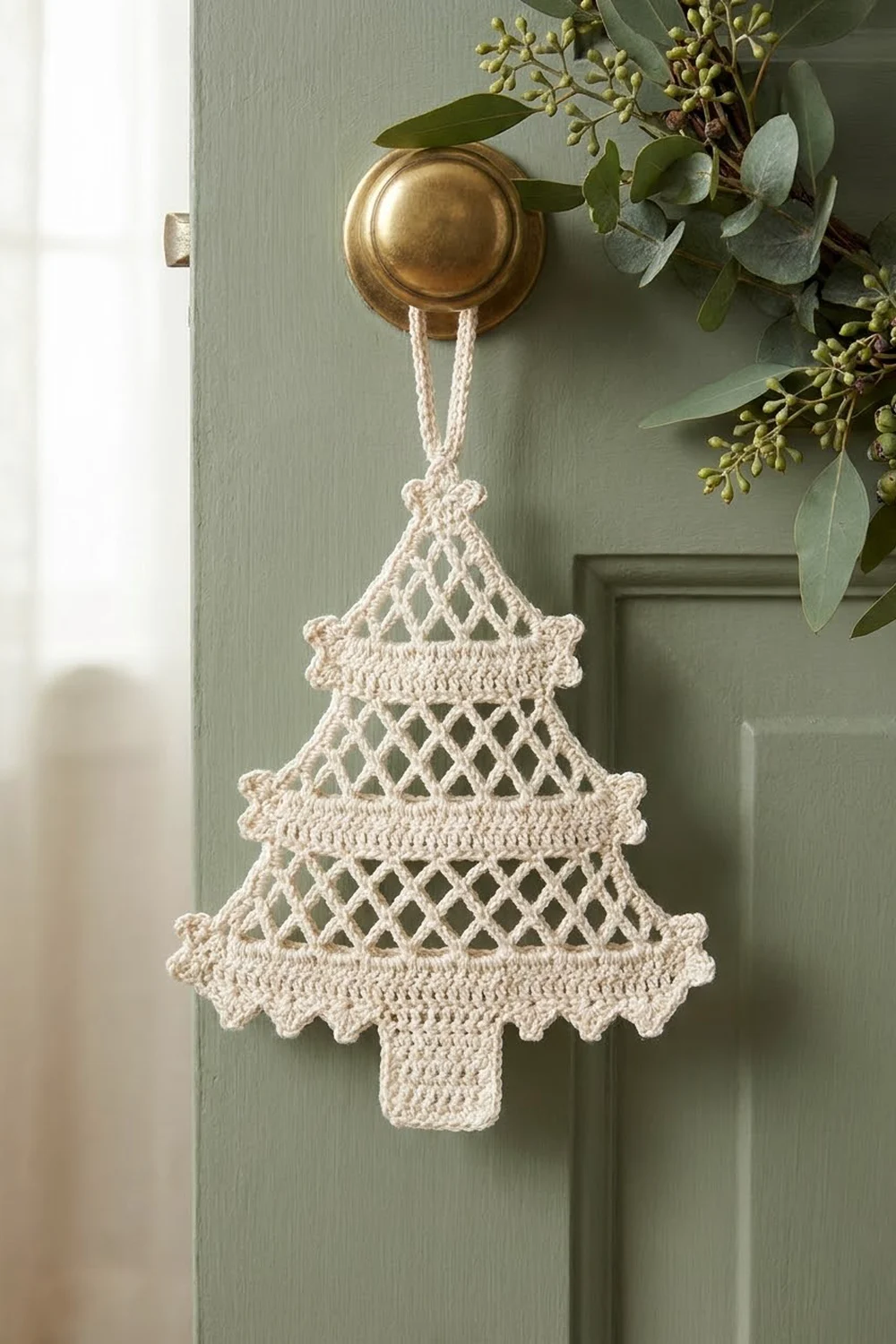 Crocheted cream Christmas tree with openwork tiered sections hangs from brass knob on sage green painted door.
