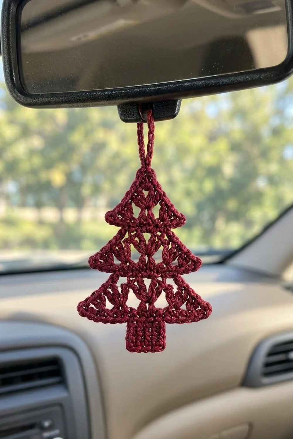 Burgundy crocheted Christmas tree ornament hanging from a car's rearview mirror with sunny landscape visible outside.