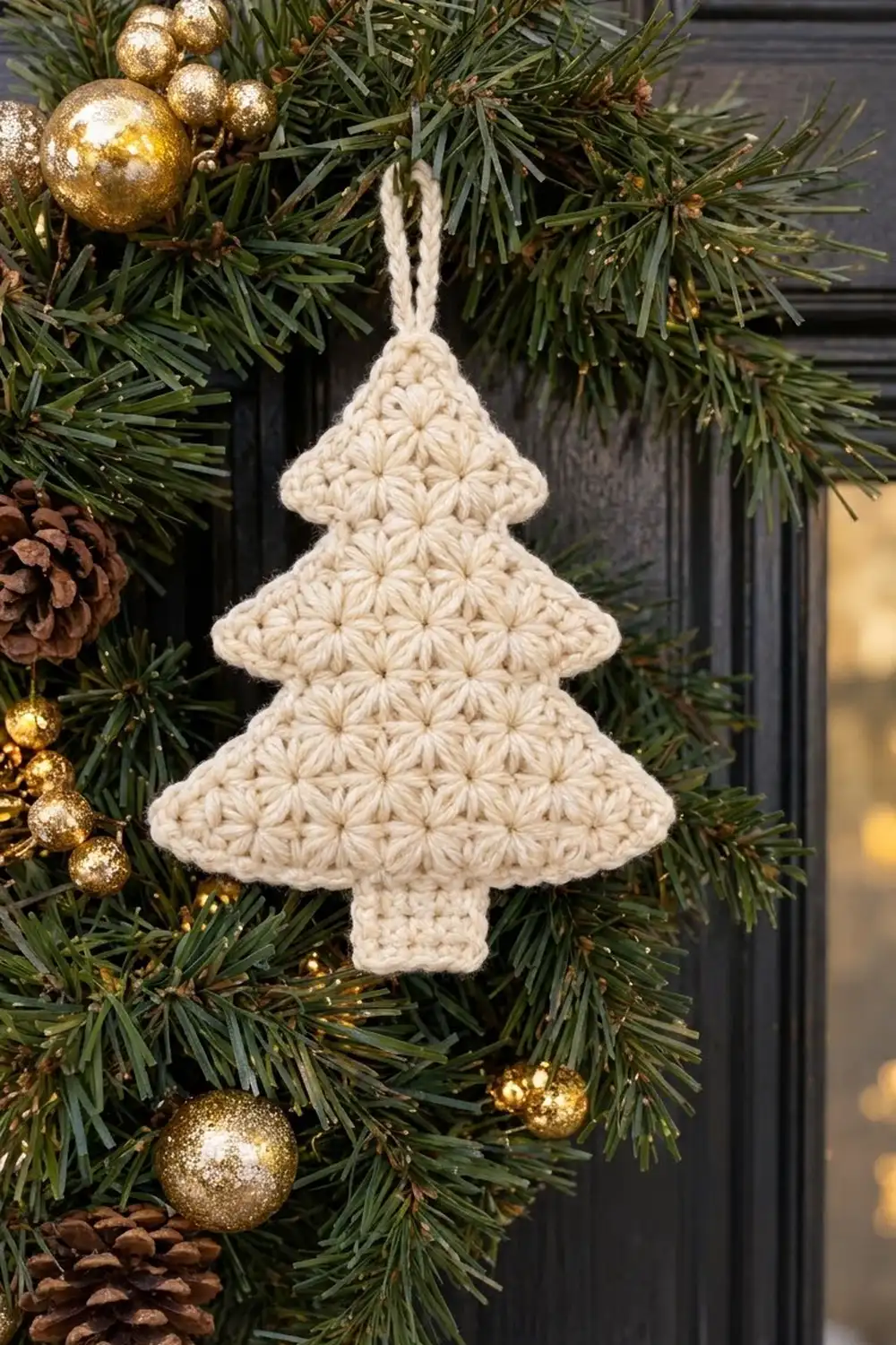 Cream crocheted Christmas tree ornament hanging among green pine garland with gold glitter baubles and brown pinecones.