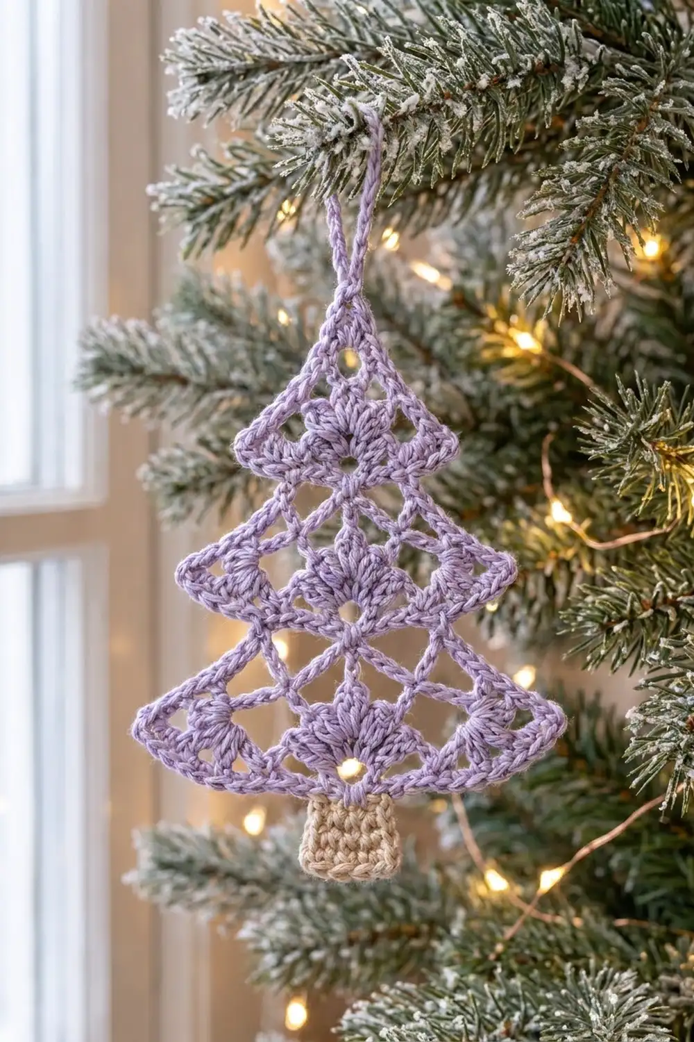Lavender crocheted Christmas tree ornament with cream trunk hanging on a frosted evergreen branch with warm white lights.
