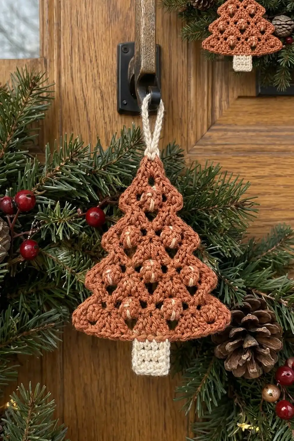Rust-colored crocheted Christmas tree ornament with cream trunk hanging on wooden door amid evergreen garland, red berries, and pinecones.
