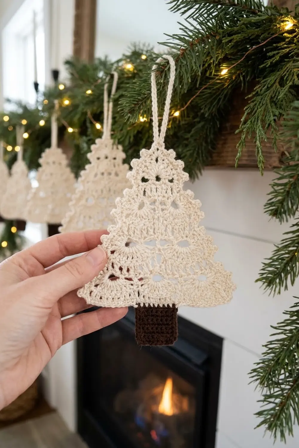 Hand holding a cream crochet Christmas tree ornament with brown trunk against twinkling holiday greenery.