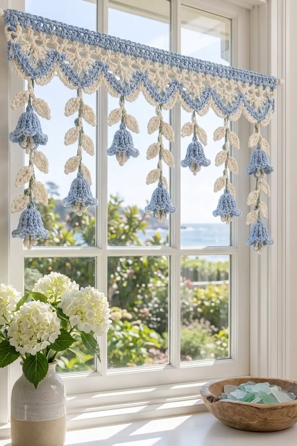 A crocheted blue and cream valance with hanging bell and leaf motifs decorates a sunny window overlooking a garden.