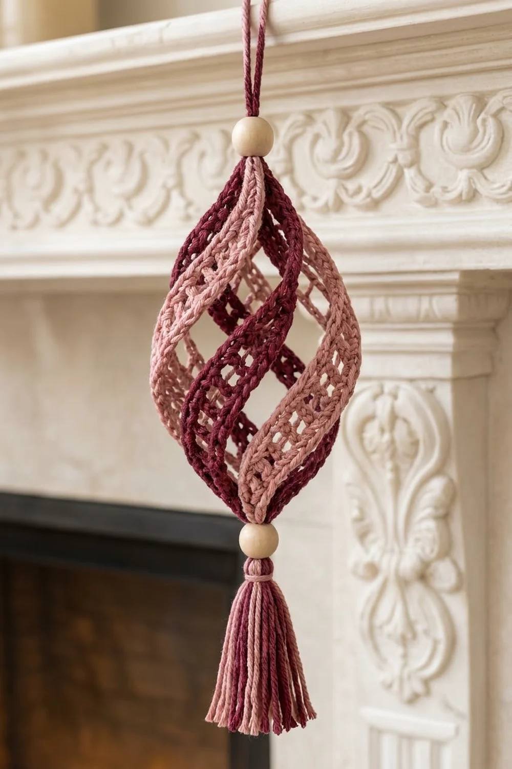 A burgundy and mauve crocheted diamond ornament with wooden beads hangs from a mantelpiece with decorative molding.