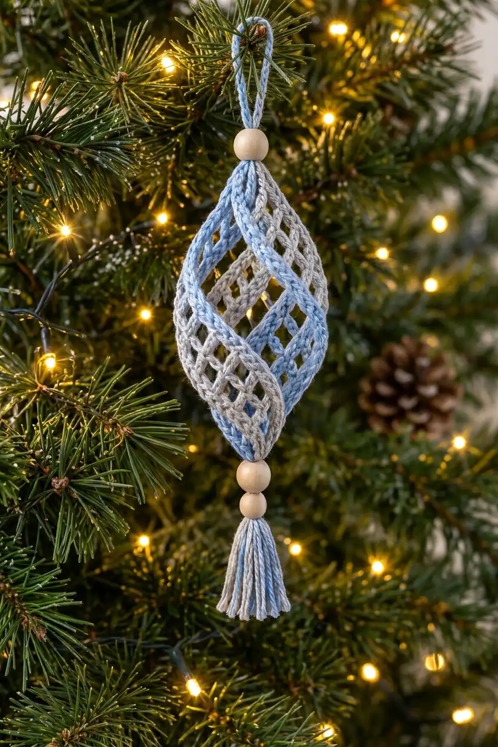 A blue and cream crocheted diamond ornament with wooden beads hangs from an evergreen branch among warm white lights.