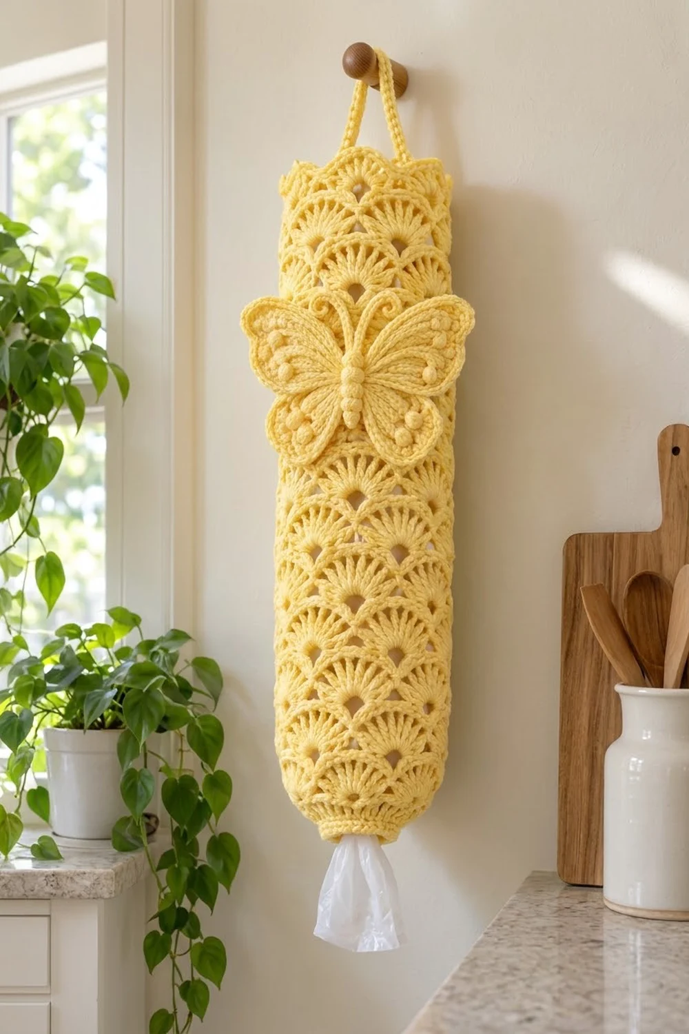 A yellow crocheted plastic bag holder with a butterfly motif hangs on a kitchen wall near plants and wooden utensils.