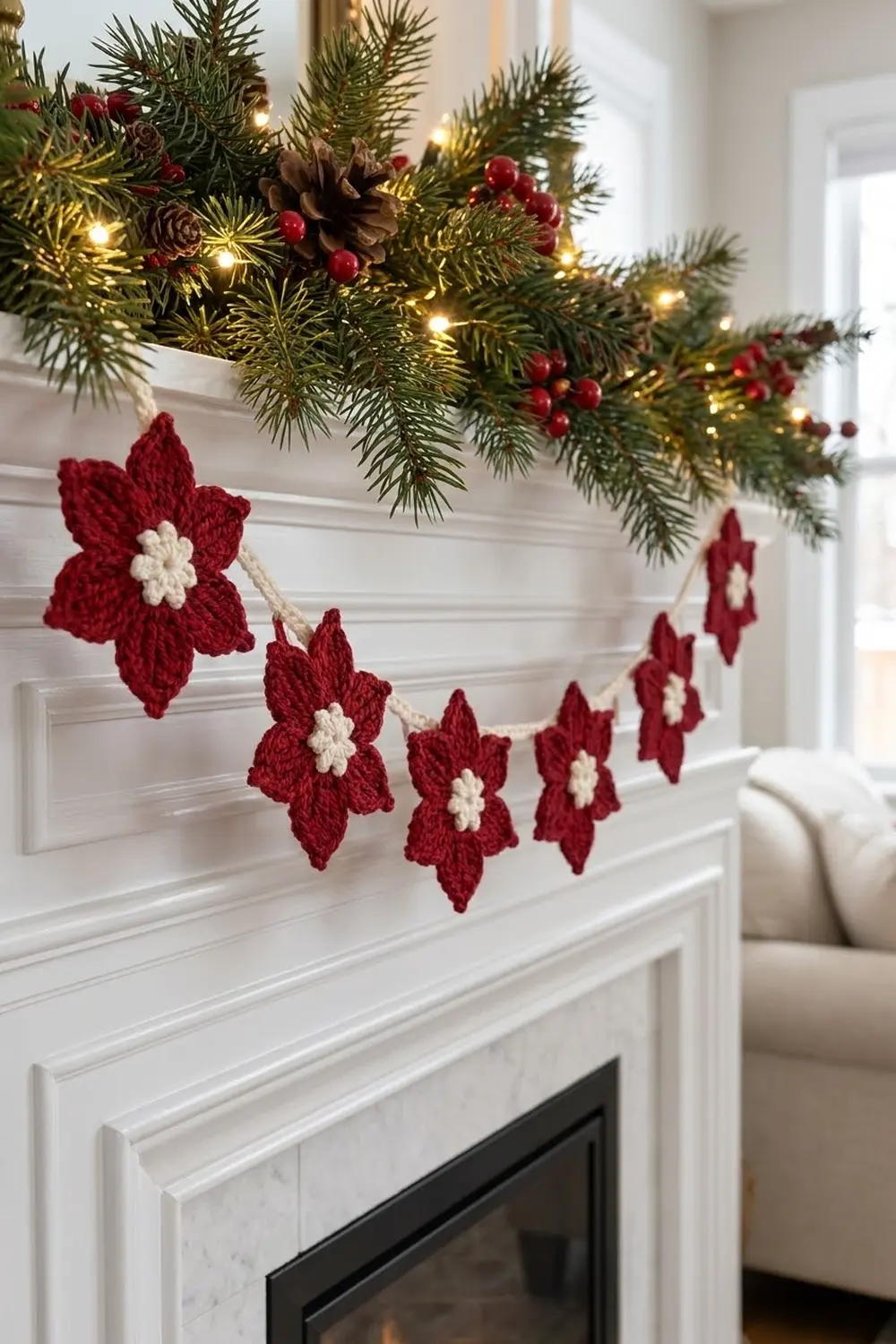 A crocheted red and white flower garland draped below a evergreen garland with lights on a white fireplace mantel.