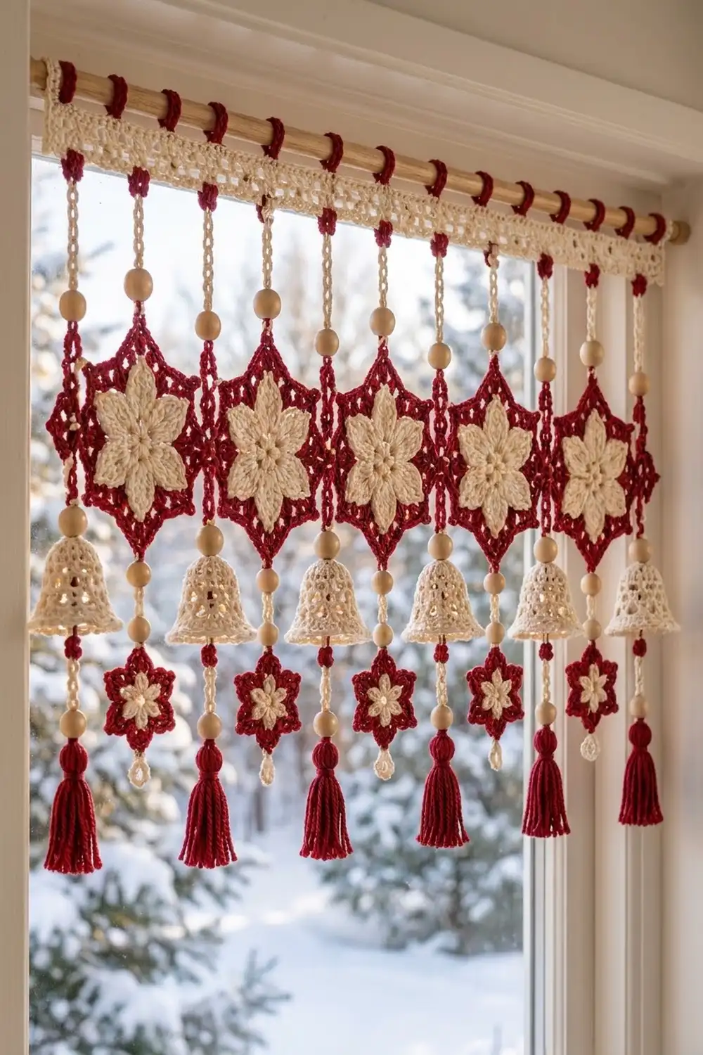 A crocheted window curtain valance featuring red and cream flowers, bells, and tassels hanging in a snowy winter window.