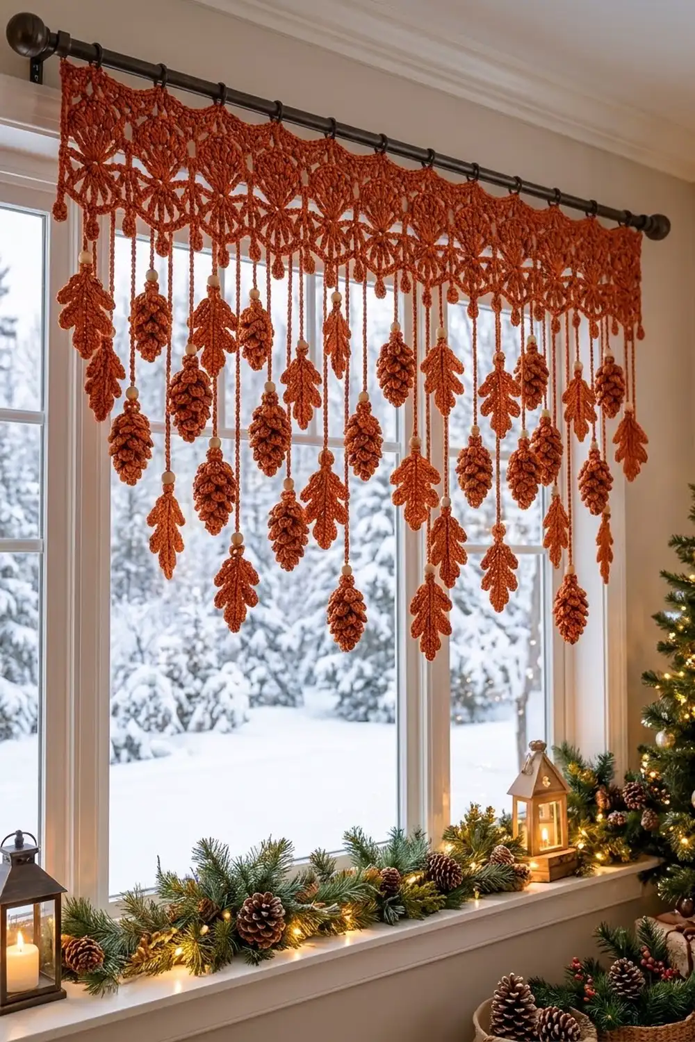Rust-colored crocheted leaf garland with hanging pinecones hangs above a winter windowsill decorated with greenery and lights.