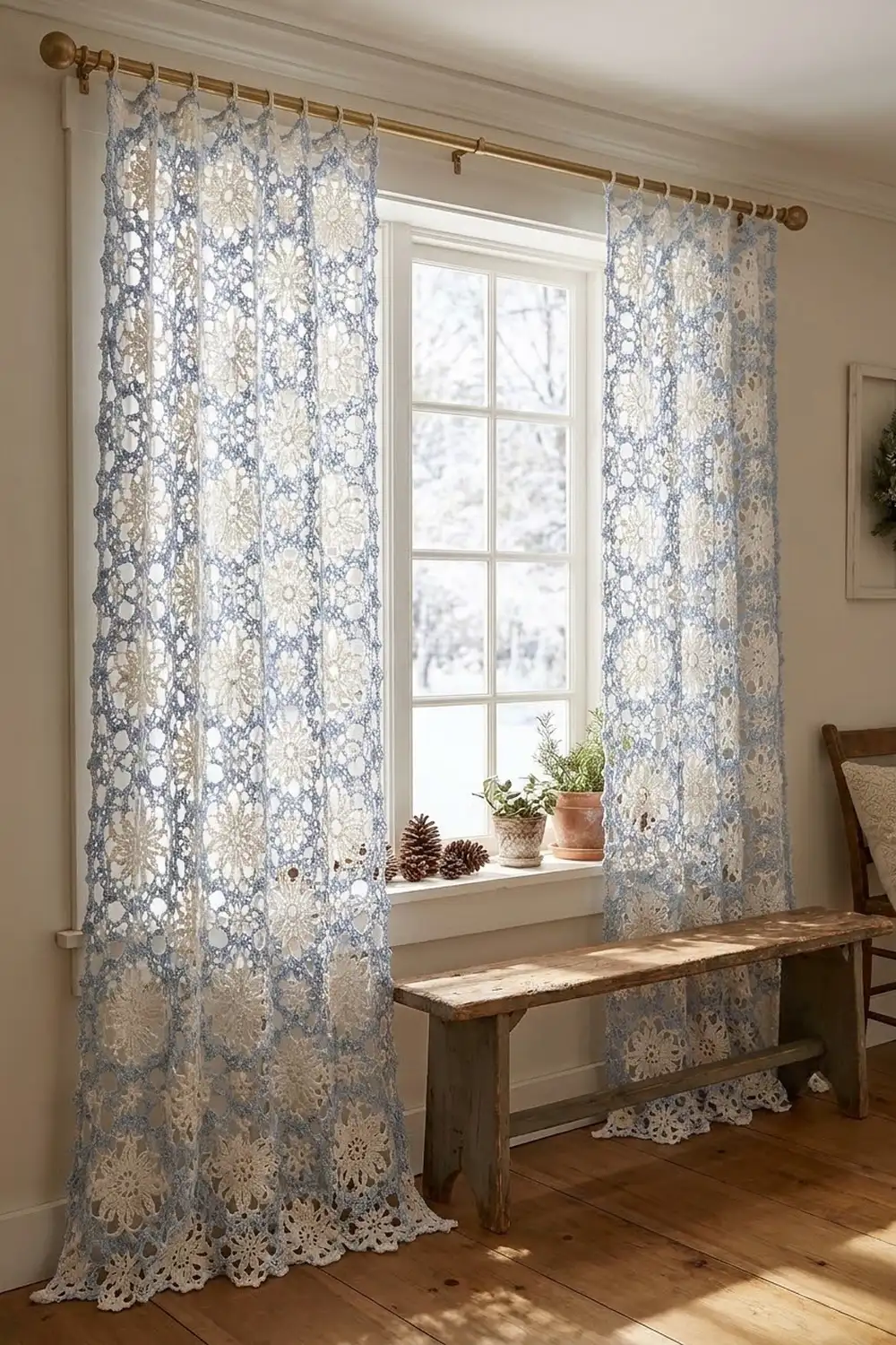 Delicate blue and cream lace curtains hang from a brass rod beside a window with potted plants and wooden bench.
