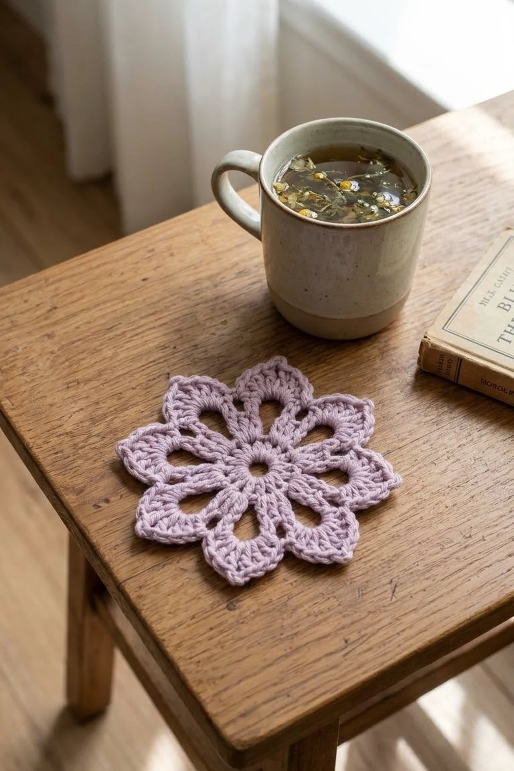 A crocheted lilac flower coaster sits on a wooden table beside a mug of tea and an open book.