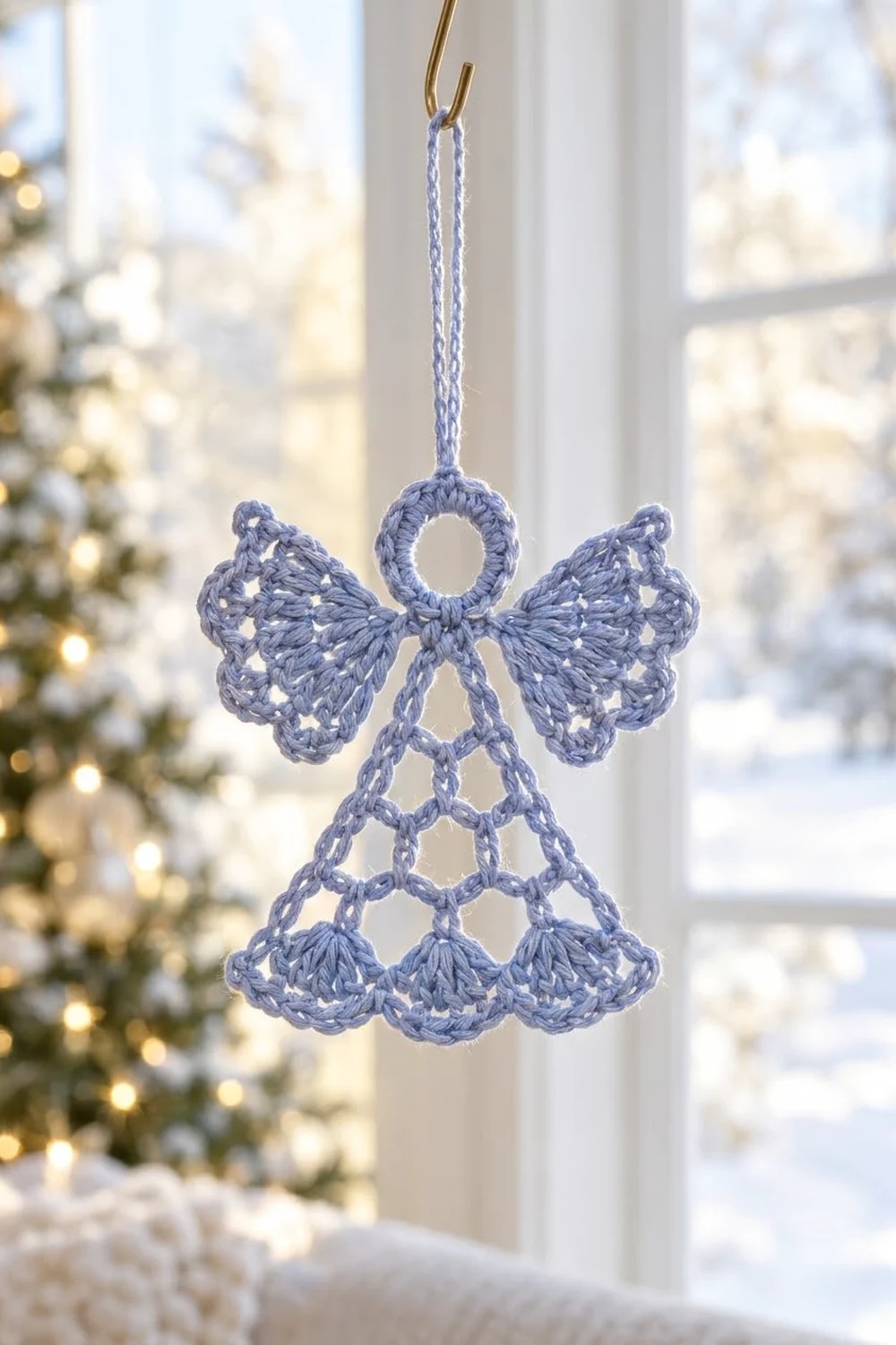 A crocheted blue angel ornament with wings hangs in a window beside a decorated Christmas tree.