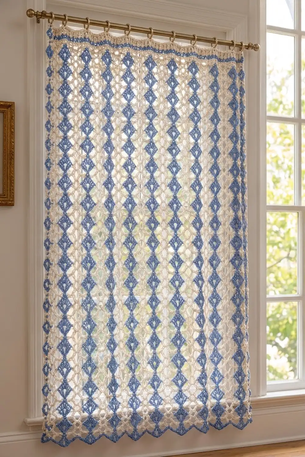 A blue and cream patterned lace curtain panel hangs from a brass rod on a tall window overlooking greenery.