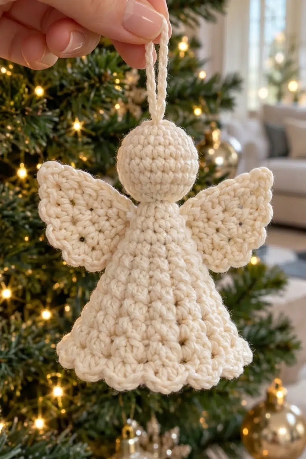 A hand holds a cream crocheted angel ornament with wings against a decorated Christmas tree with golden lights.