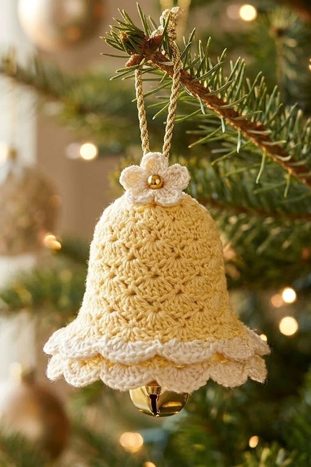 A cream and gold crocheted bell ornament with a flower detail hangs from an evergreen branch near twinkling lights.