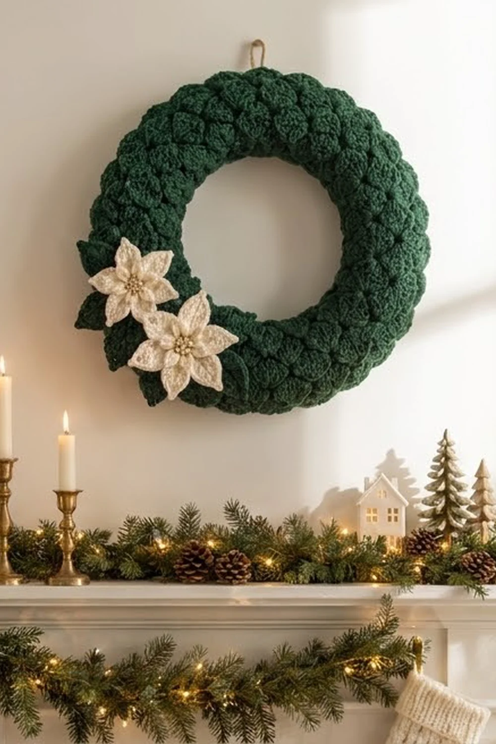 A green knitted wreath with cream flowers hangs above a mantelpiece decorated with garland, candles, and miniature houses.