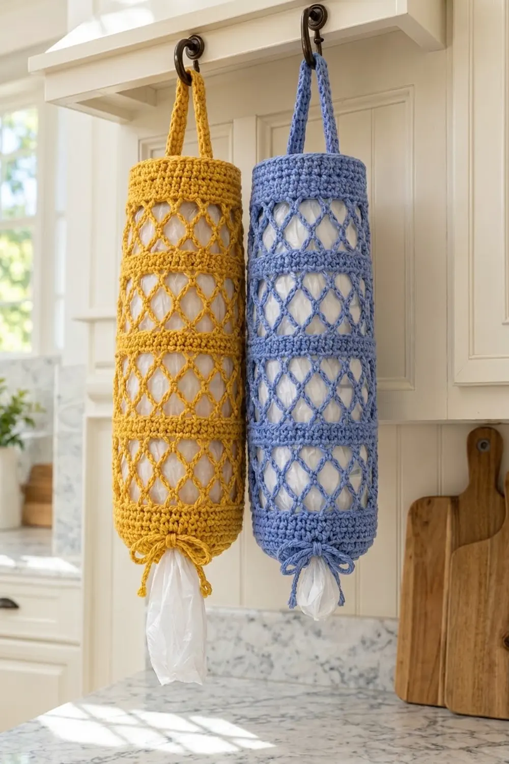 Two crocheted plastic bag holders in yellow and blue hang from ornate hooks in a bright kitchen.