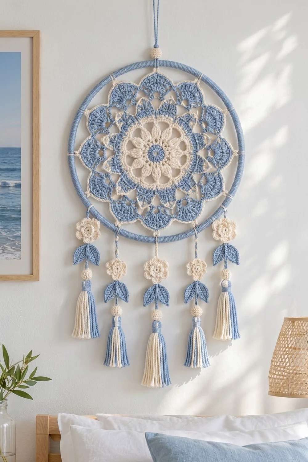 A blue and cream crocheted dreamcatcher with floral motifs and tassel dangles hangs on a white wall above a bed.