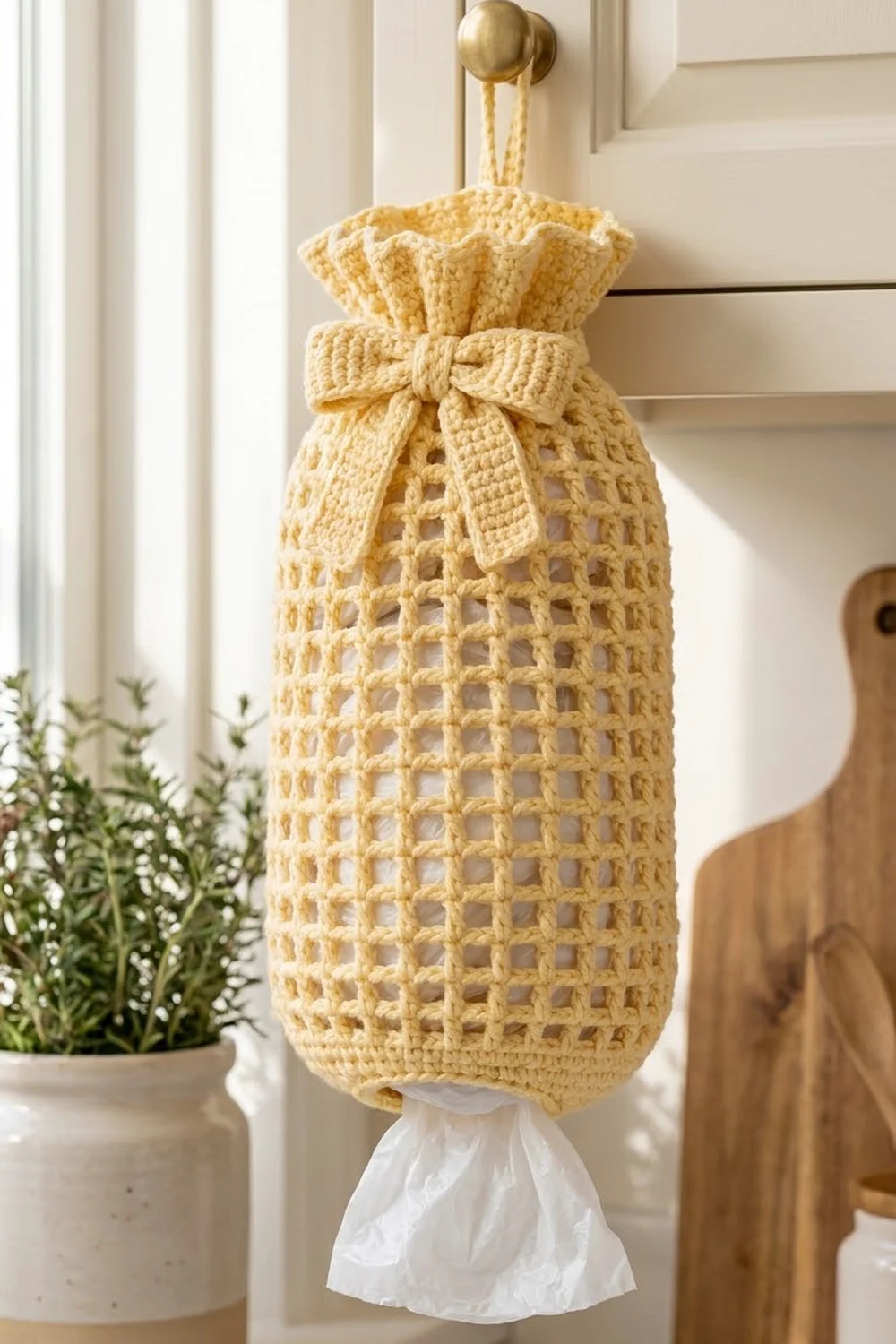 A cream-colored crocheted plastic bag holder with a bow hangs on a brass hook in a bright kitchen.