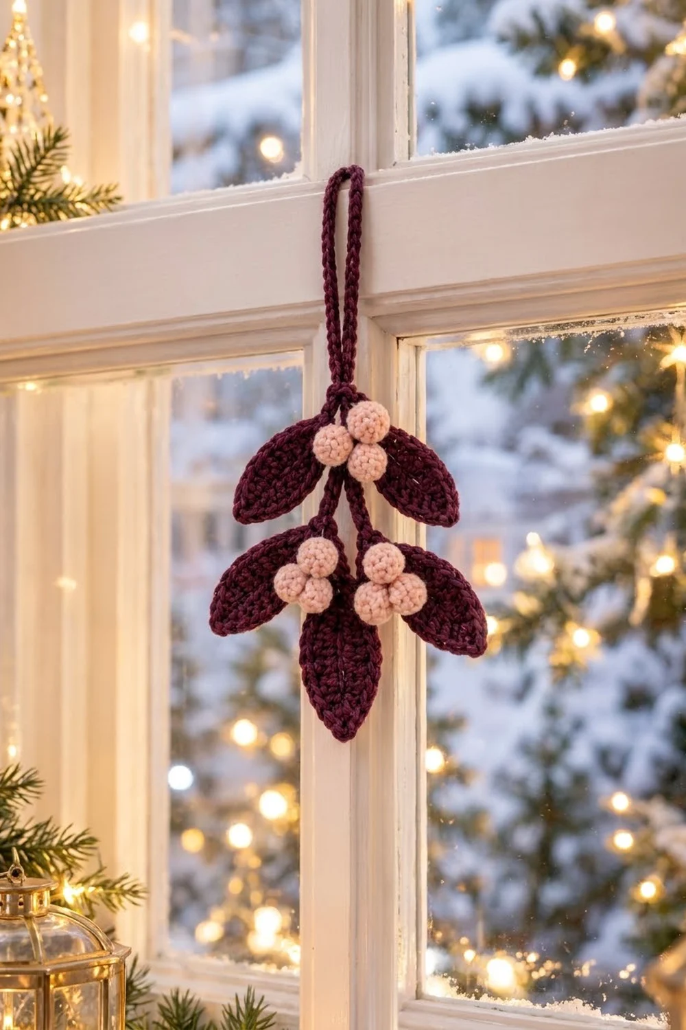 A crocheted burgundy mistletoe ornament with cream berries hangs from a window frame in snowy holiday decor.