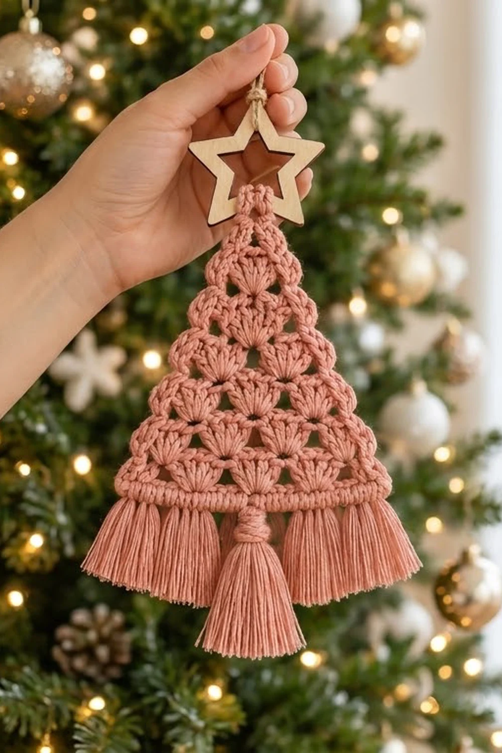 A hand holds a dusty rose macramé Christmas tree ornament with a wooden star topper against a decorated evergreen tree.