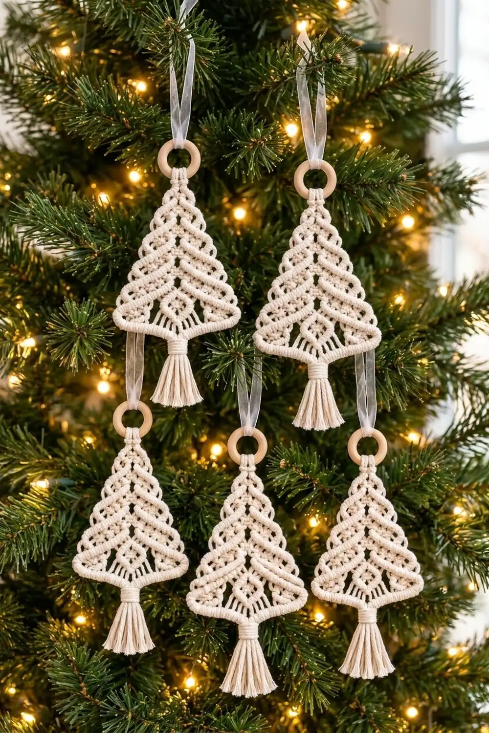 Five cream macramé Christmas tree ornaments with tassels hanging from gray ribbons on a lit evergreen tree.