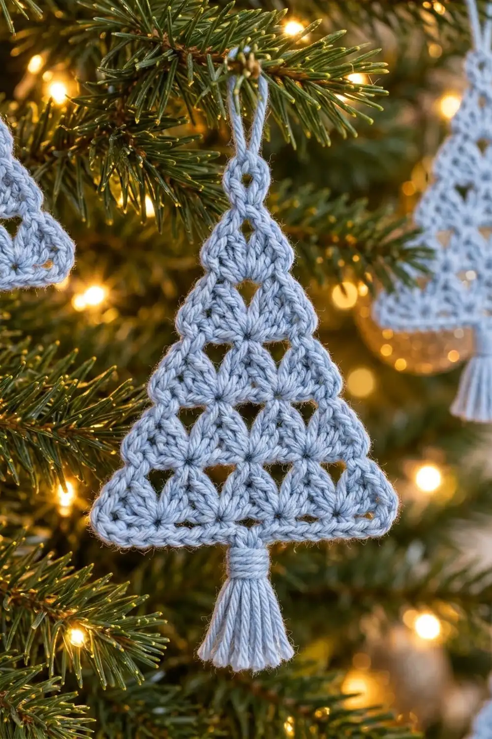 A light blue crocheted Christmas tree ornament with openwork pattern hangs from an evergreen branch among warm white lights.