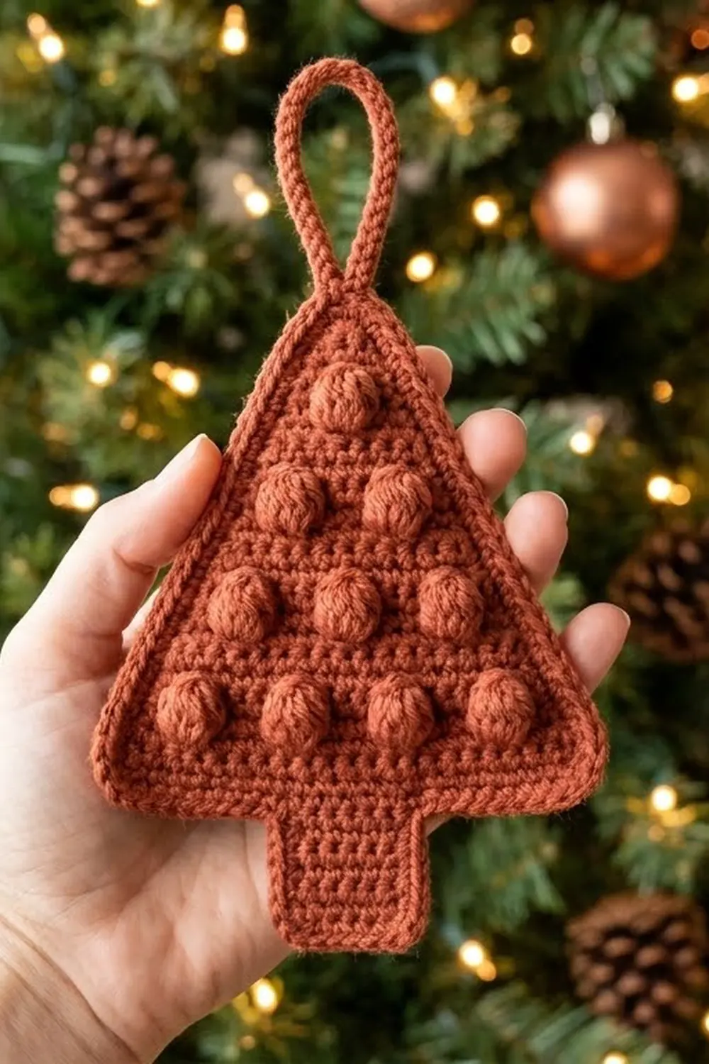 A hand holding a terracotta crocheted Christmas tree ornament against a decorated holiday tree backdrop.